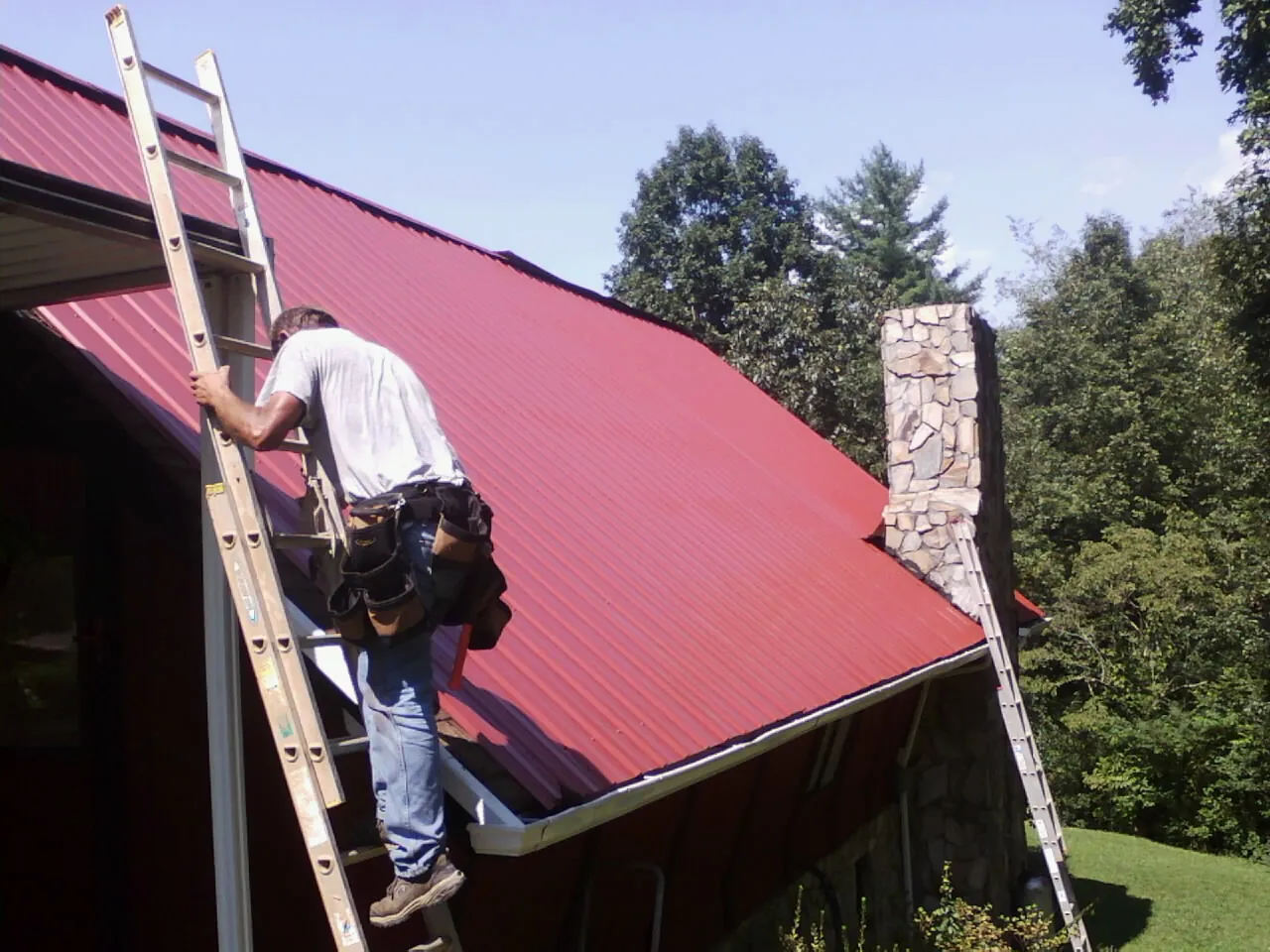 Roofer on a ladder beside a red metal roof and stone chimney — Soffit & Fascia Repair by Roofing Brothers in Knightstown